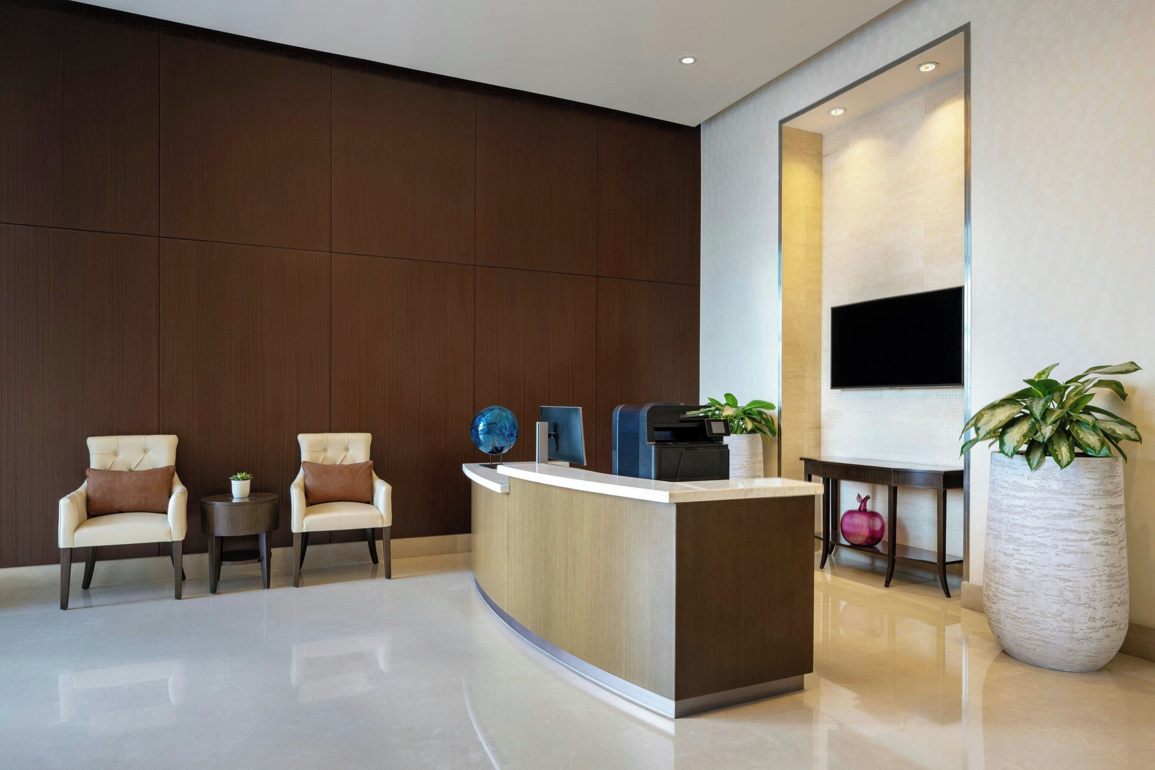 A modern reception area with a curved reception desk, two cushioned chairs, a small table with a globe and a plant, and a large potted plant.
