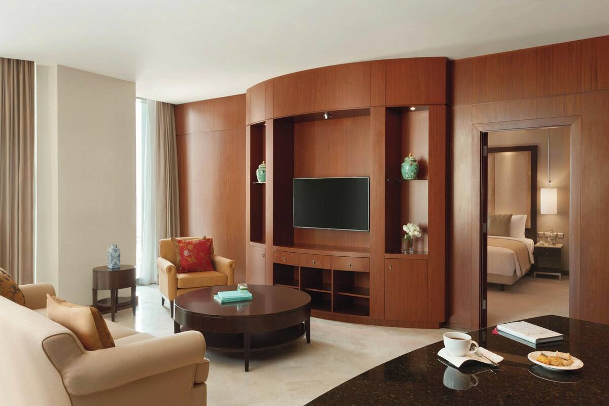 A modern hotel room with a curved wooden panel wall, a flat-screen TV, a round coffee table, a sofa, and a bed with a nightstand.