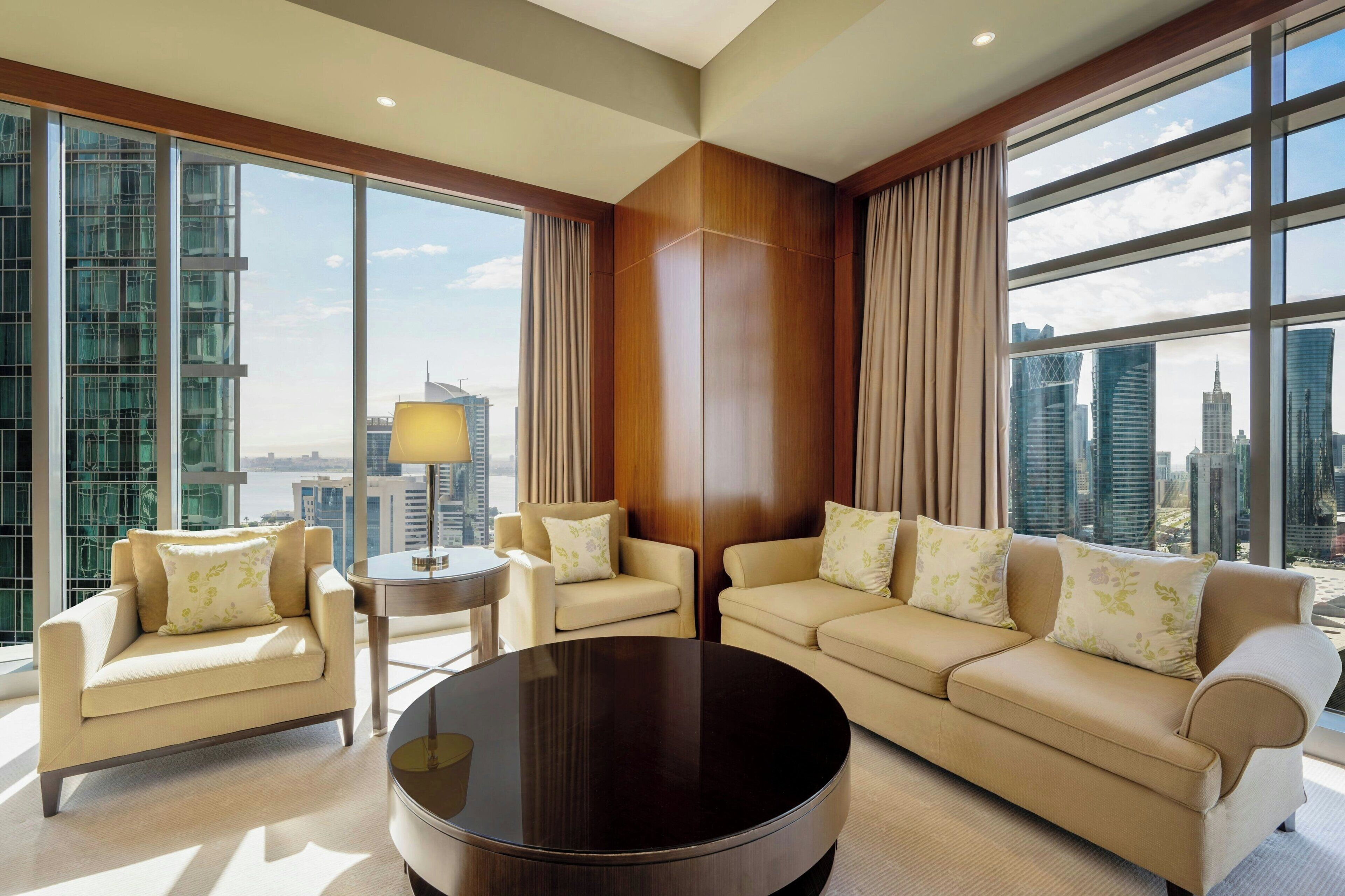 A modern hotel room with a large window offering a city view, a round coffee table, two sofas, and a small side table.
