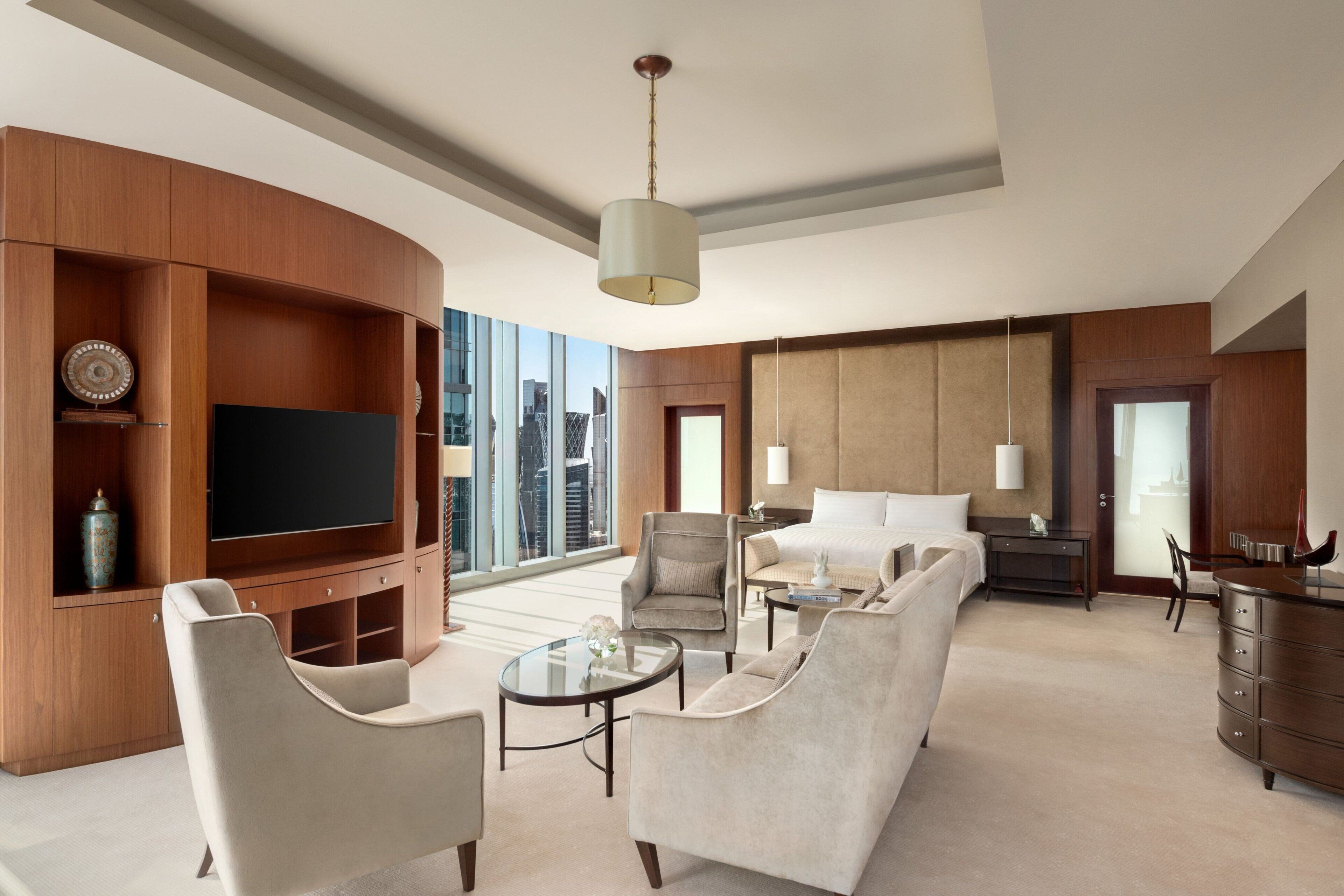 A modern hotel room with a large TV, comfortable seating, a bed, and a view of the city.