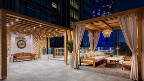 Un área de lounge en la azotea con una pérgola de madera, un sofá, una mesa y una planta grande en maceta.