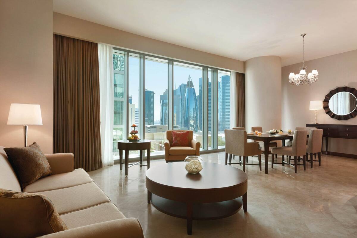 A hotel room with a large window offering a city view, a round dining table, a sofa, and a chandelier.