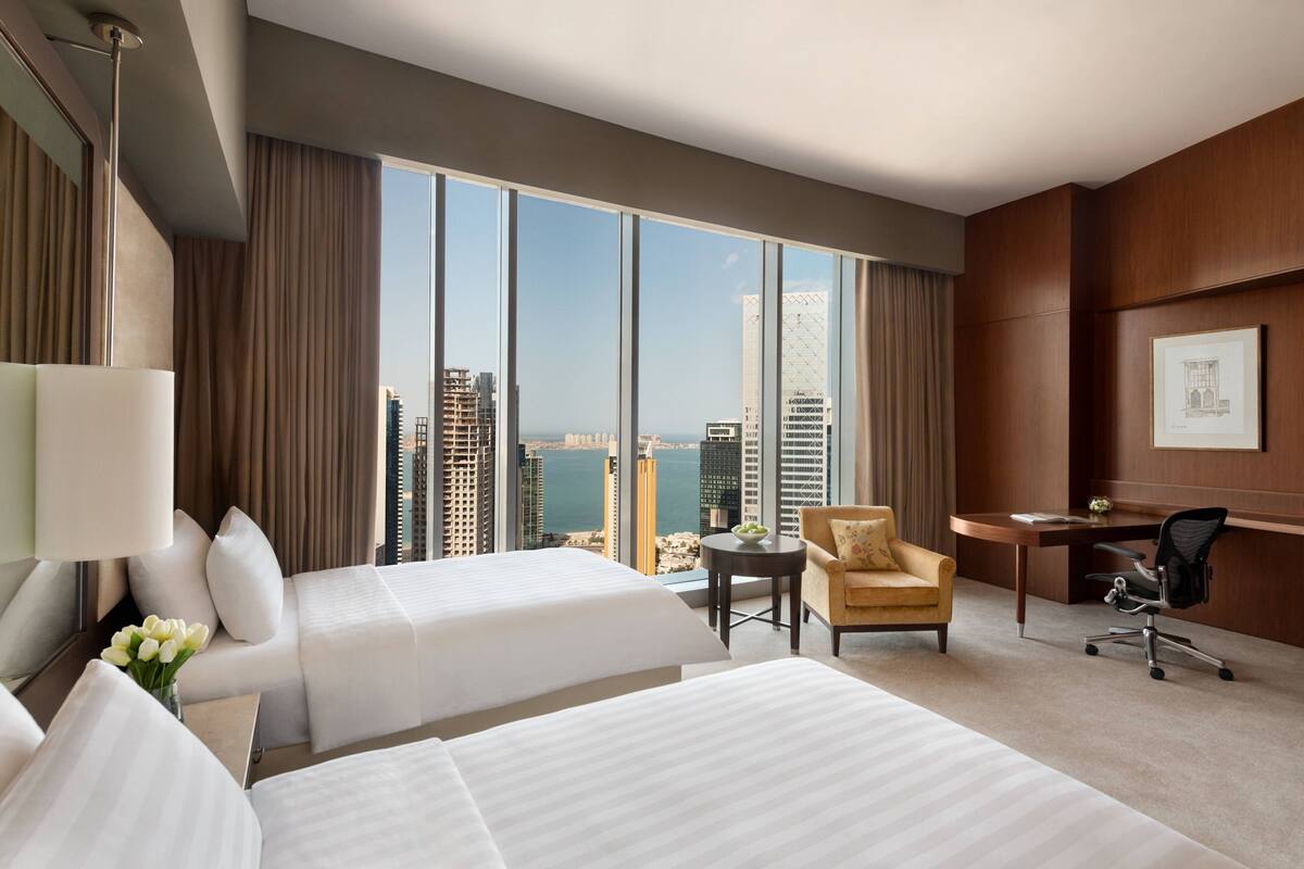 A hotel room with two beds, a desk, a chair, and a large window offering a city view.