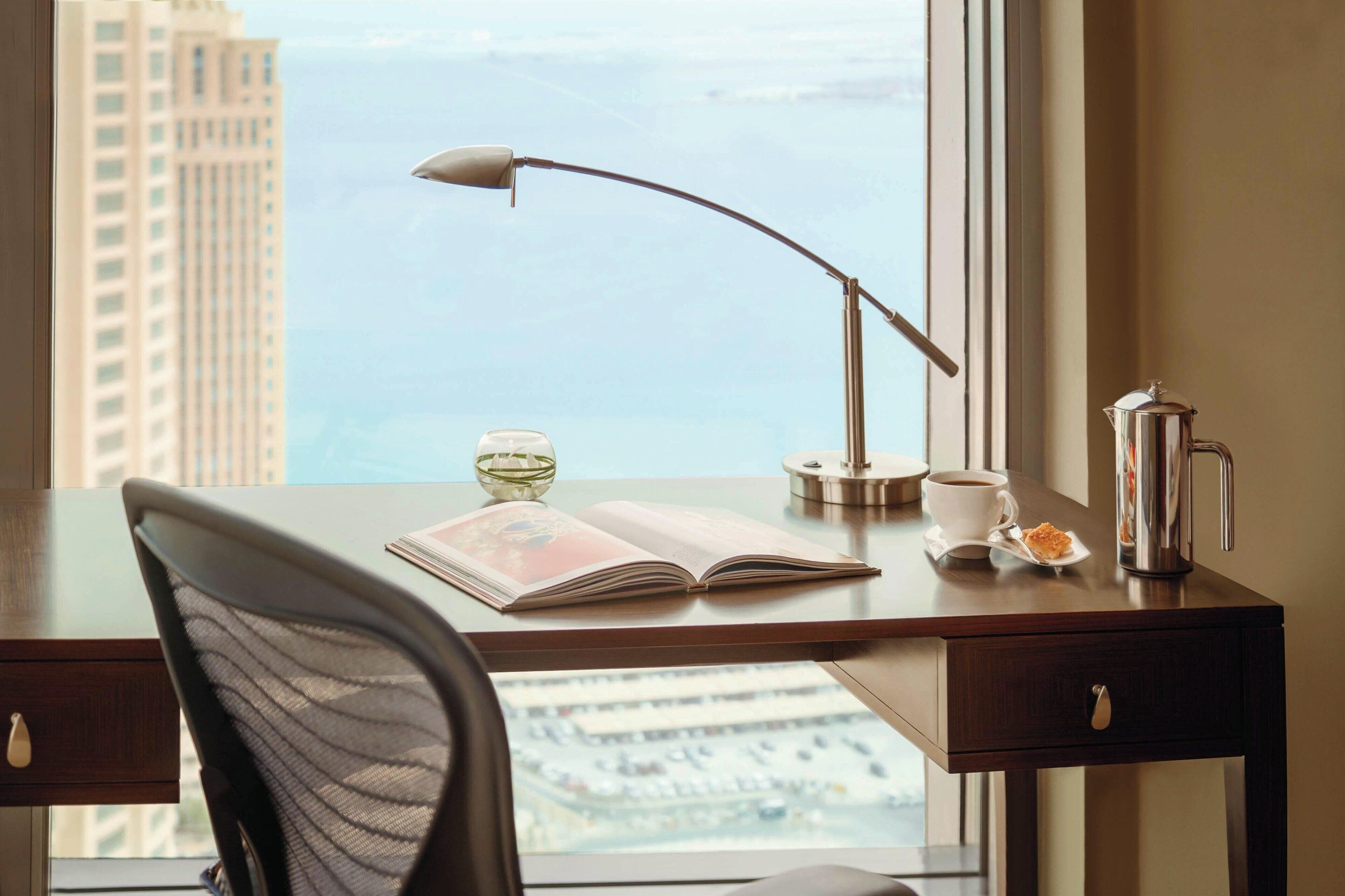 Un escritorio con un libro, una taza y una cafetera al lado de una ventana que da a vistas de la ciudad.