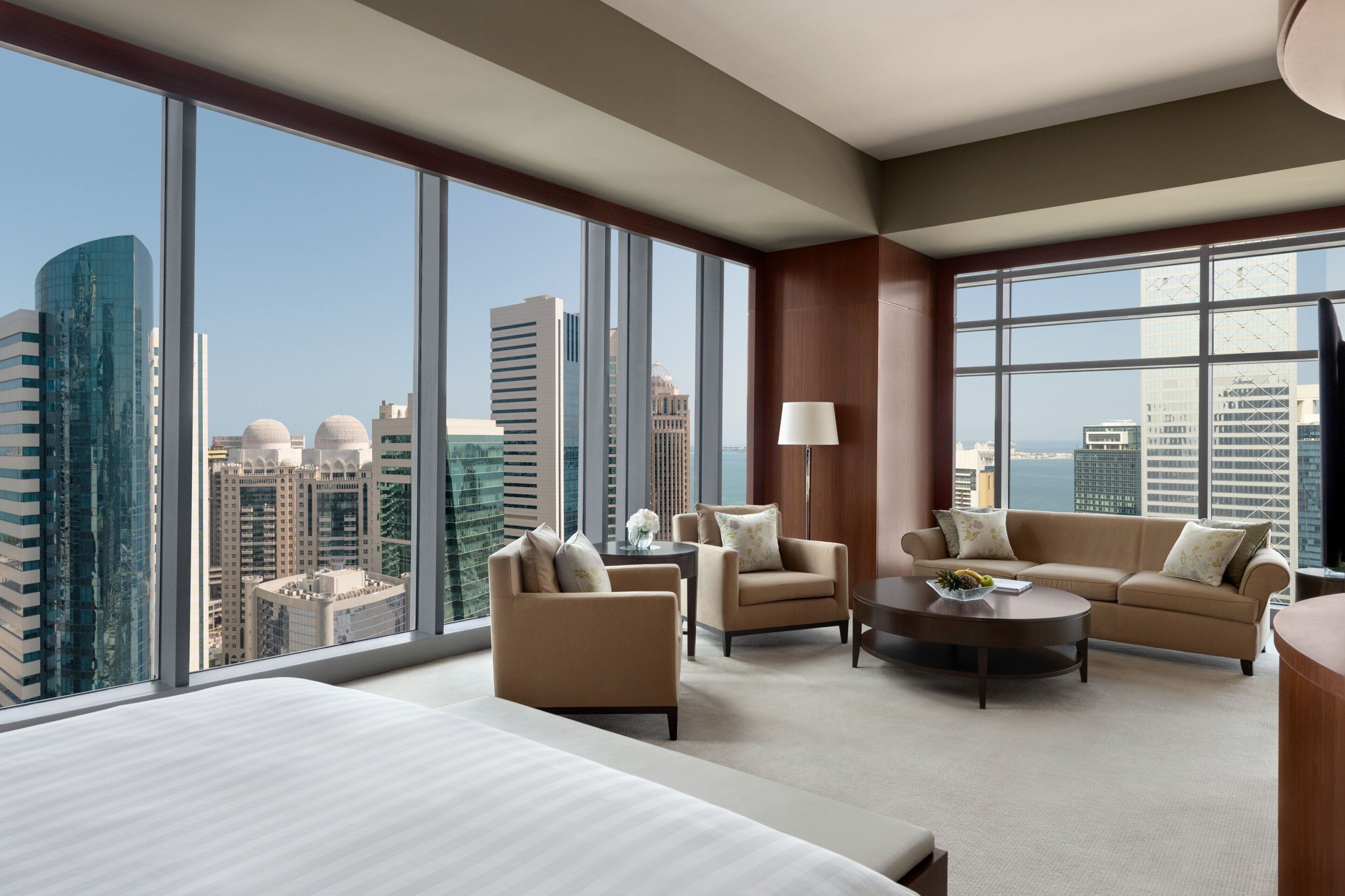 Een moderne hotelkamer met een groot bed, twee fauteuils, een bank, een salontafel en uitzicht op de skyline van de stad door middel van kamerhoge ramen.