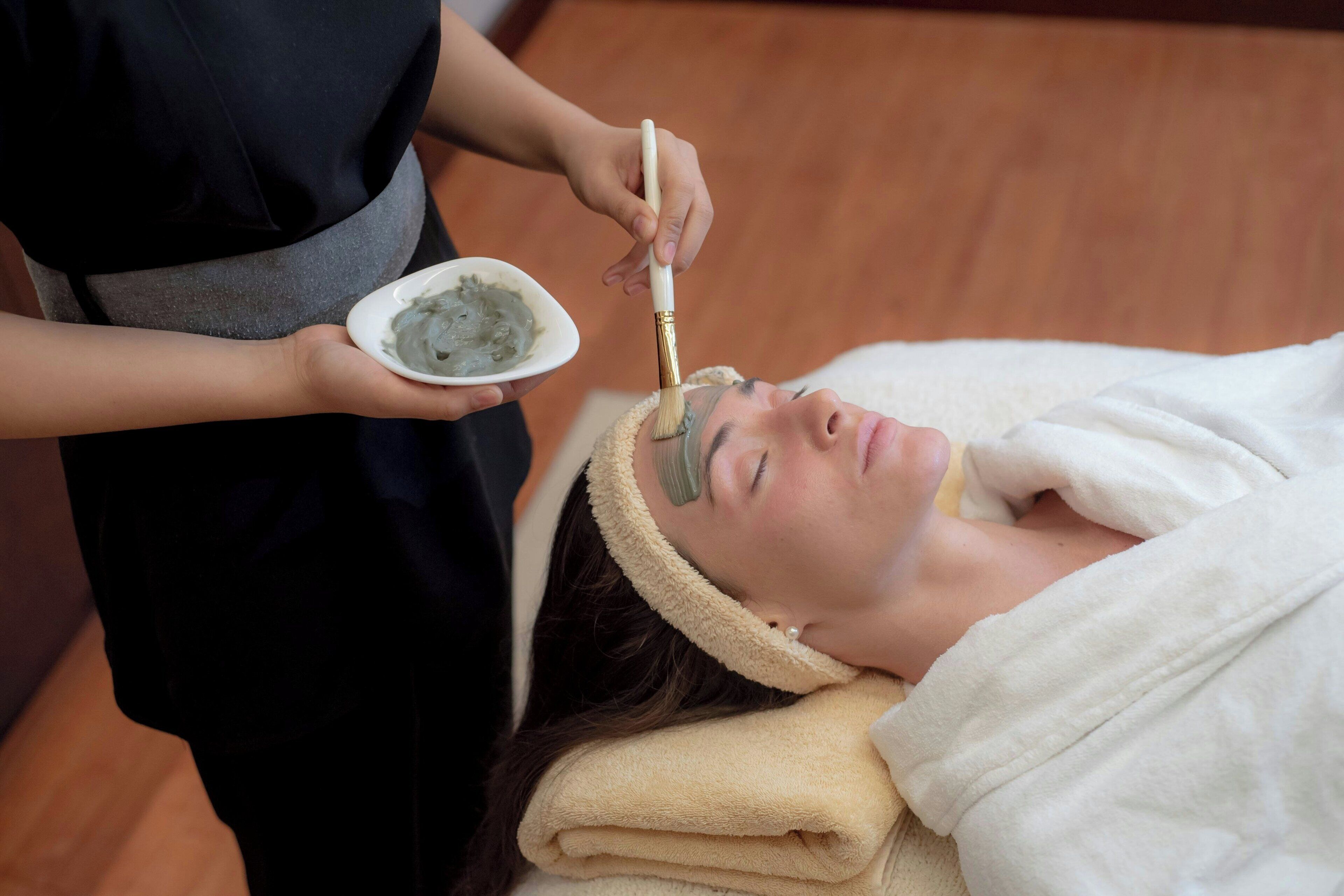 A person receiving a facial treatment with a brush, in a room with wooden flooring.