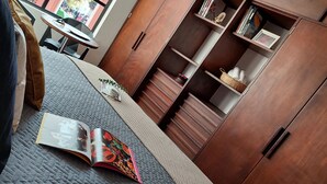 Een kamer met een bed, een klein tafeltje, een boekenkast en een raam met uitzicht op mensen buiten.