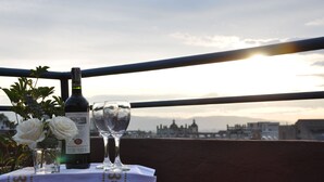 Een fles wijn en twee glaasjes op een tafel met uitzicht op een stadslandschap bij zonsondergang.