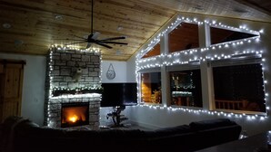 A cozy living room with a stone fireplace, a ceiling fan, and a large window with string lights. - Custom built cabin sitting on several acres lot in Pinetop-Lakeside! (Pinetop-Lakeside)