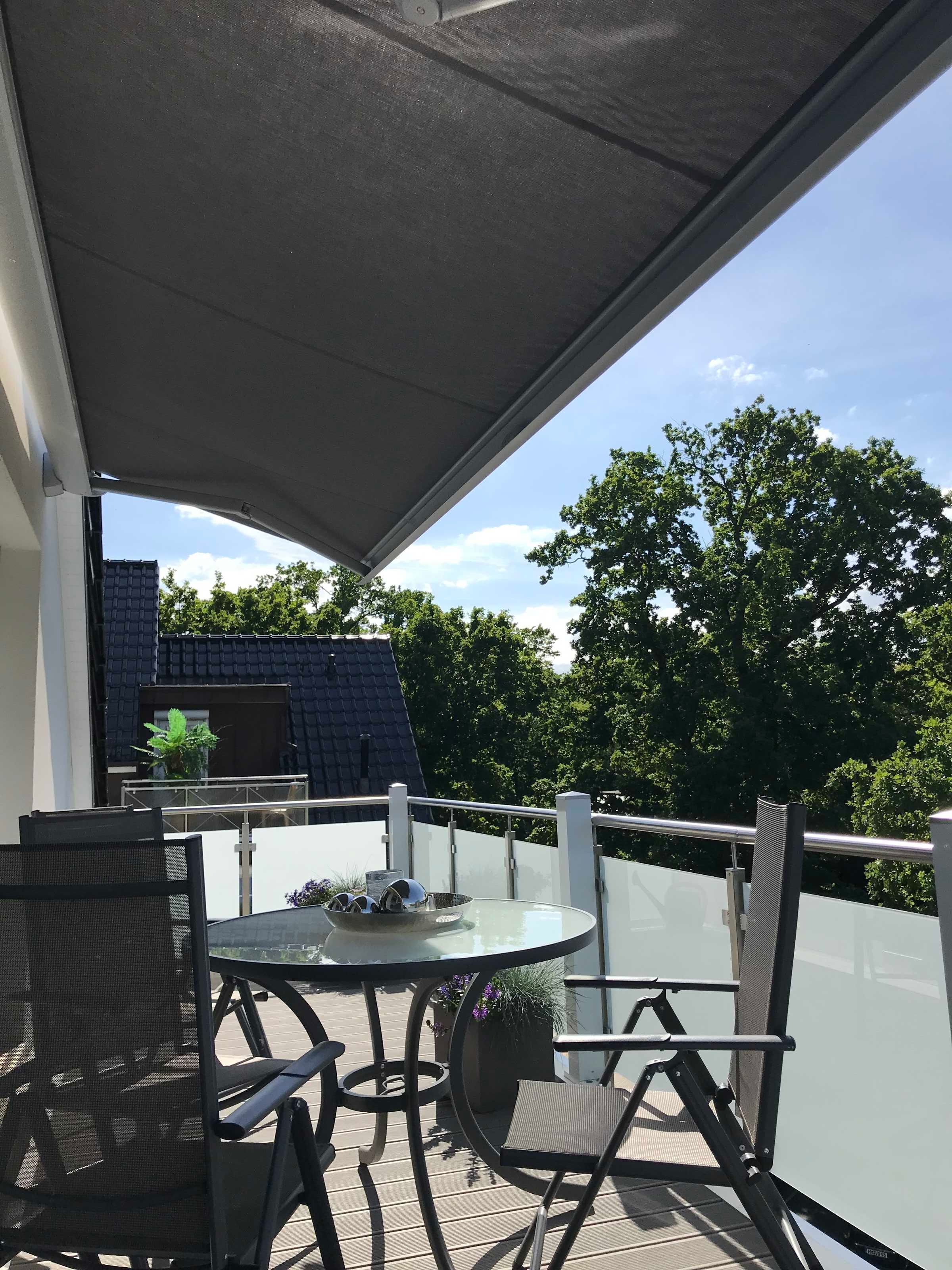 Ein überdachter Außenbereich mit Tisch und Stühlen, einer Glasbrüstung und Blick auf Bäume sowie ein Gebäude im Hintergrund.