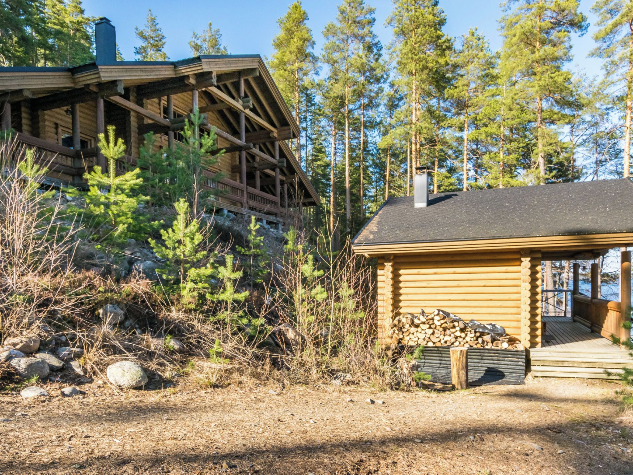 A cabin with a wooden deck and a covered porch, surrounded by trees and a rocky ground.
