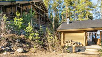 A cabin with a wooden deck and a covered porch, surrounded by trees and a rocky ground.