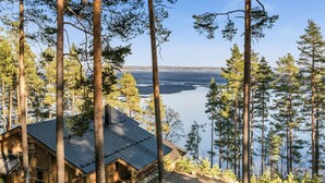 A cabin with a sloped roof, surrounded by tall pine trees, overlooking a calm lake.