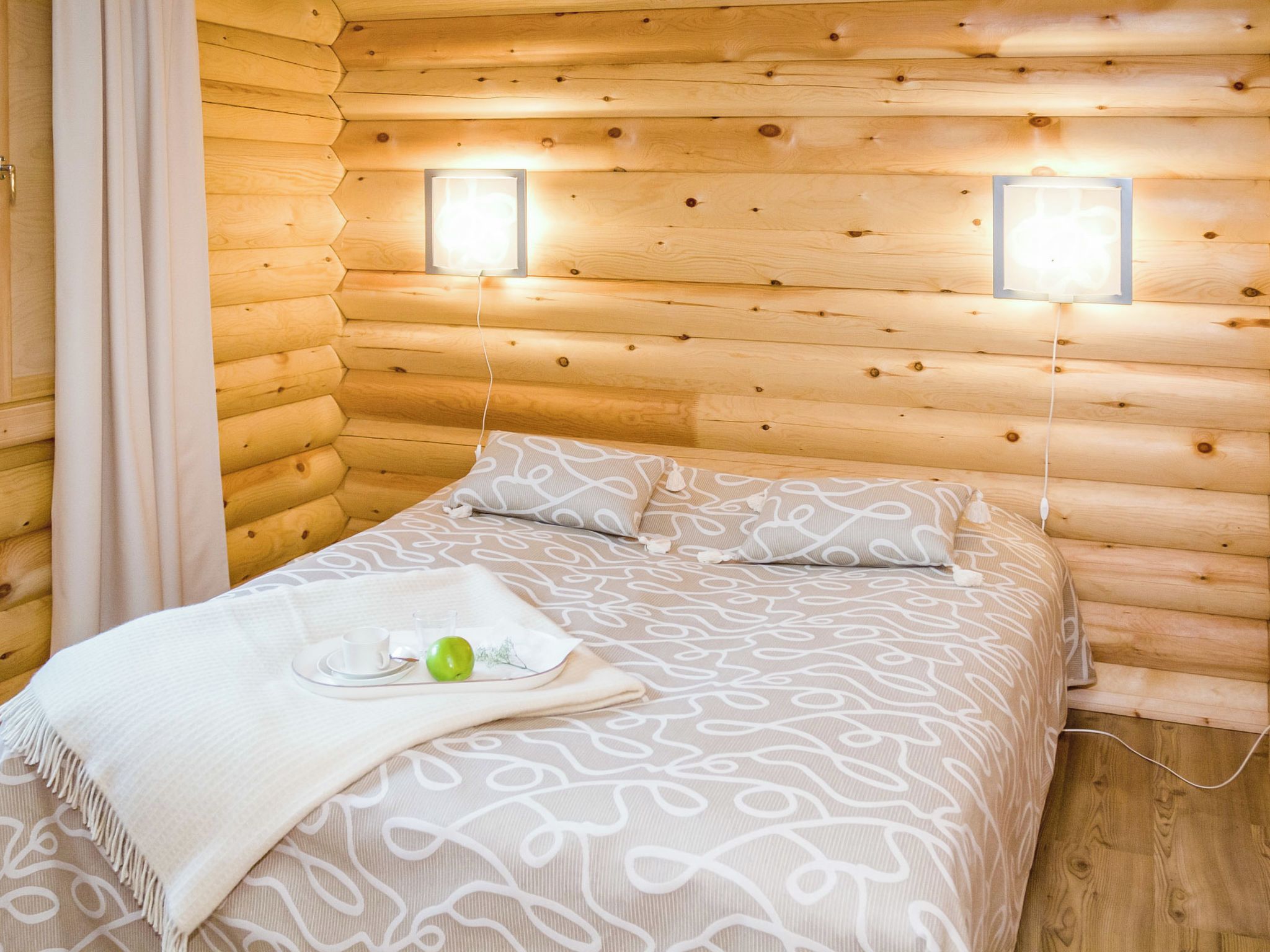 A log cabin bedroom with a bed, pillows, a bedside table with a cup and apple, and two wall-mounted lights.