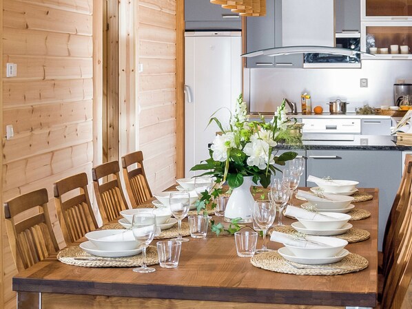 Une table à manger dressée pour un repas, avec des chaises, un vase de fleurs et une cuisine en arrière-plan.
