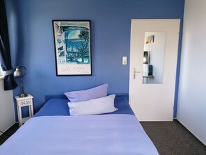 Een slaapkamer met een bed, nachtkastje, lamp, deur en een kunstwerk aan de muur.