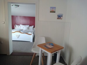 A bedroom with a bed, a chair, a small table, and a door leading to another room.