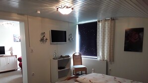 A bedroom with a bed, a chair, a desk, a TV, and a window with curtains. - Apartment - Hotel Möven - Kieker (Friedrichskoog)