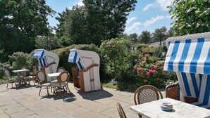 An outdoor patio with wicker furniture and striped umbrellas, surrounded by greenery and flowers. - Apartment - Hotel Möven - Kieker (Friedrichskoog)