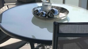 Une table ronde en verre, sur laquelle repose un plateau en métal portant trois verres et une bougie, est entourée de chaises en osier.