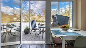 A modern dining area with a glass door leading to an outdoor deck, a table with chairs, and a bowl of fruit on the table. - Silbermoewe - Nordic Wave Apartments (Timmendorfer Strand)