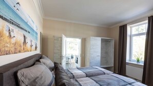 A bedroom with a large bed, a painting of a beach scene, a wardrobe, and a window with curtains. - Silbermoewe - Nordic Wave Apartments (Timmendorfer Strand)