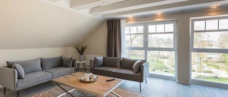 A modern living room with a grey sectional sofa, a wooden coffee table, and a large window offering a view of a garden.