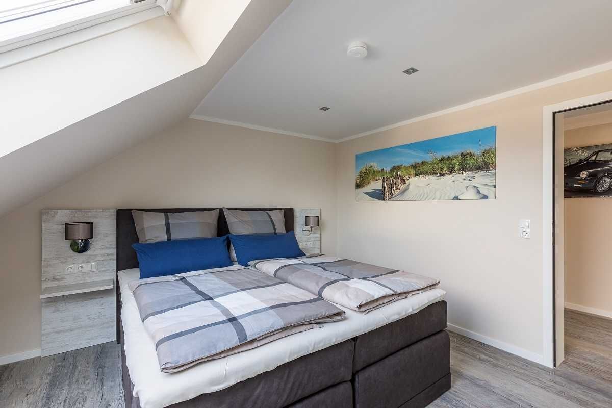 Un dormitorio con techo inclinado, una cama con colcha a cuadros grises y blancos, dos almohadas azules, cabecera con lámpara, lámpara de pared y un cuadro de una playa.