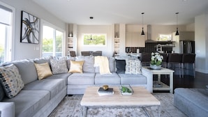 Ein modernes Wohnzimmer mit grauem Sofa, einem Couchtisch und einem Essbereich, im Hintergrund ist eine Küche erkennbar.