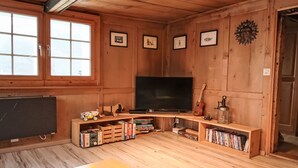 A cozy room with wooden walls, a large flat-screen TV, wooden shelves with books, and a window with blinds. - Feu de Joie by Interhome (Segnas)