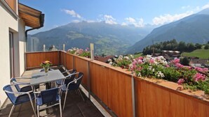 Un balcon avec une table et des chaises, donnant sur un paysage montagneux parsemé de maisons et de verdure.