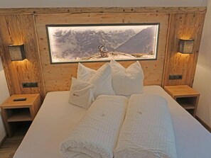 Une chambre à coucher avec une tête de lit en bois, un grand tableau encadré représentant un paysage de montagne, du linge de lit blanc et deux tables de chevet avec des lampes.