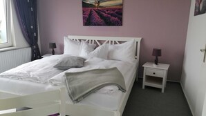 Un dormitorio con una cama blanca, dos mesitas de noche con lámparas, una ventana con cortinas y un cuadro de un campo de lavanda en la pared.