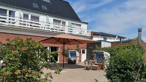 Una casa de dos pisos con un balcón, un patio con mobiliario exterior y un jardín con árboles frutales.