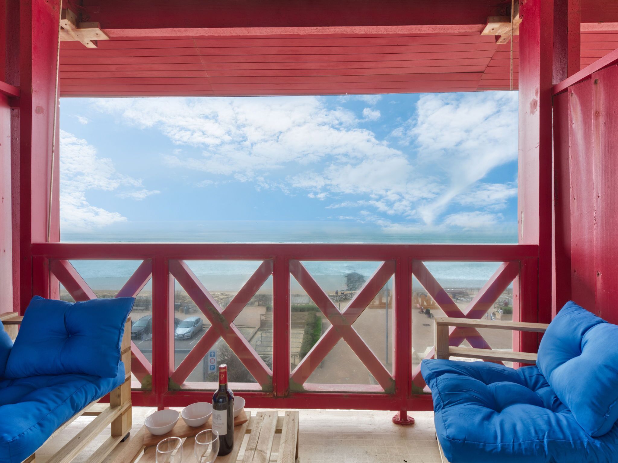 Ein Balkon mit roten Geländern, zwei blaue Sitzgelegenheiten mit Polstern, ein Holztisch mit einer Weinflasche und Gläsern sowie Blick auf das Meer und den Strand.