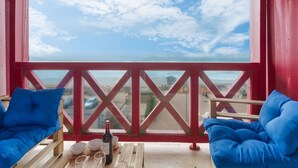 Un balcón con barandillas rojas, dos sillas con cojines azules, una mesa de madera con una botella de vino y copas, y vista al océano y a la playa.