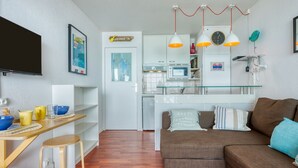 Una sala de estar moderna con un sofá marrón, una mesa de comedor de madera con taburetes, una zona de cocina con armarios blancos y un televisor montado en la pared.