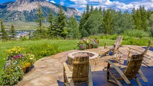 A patio with wooden chairs and a fire pit, surrounded by flower beds and a mountainous backdrop. - Sleeps 23, 8 BR w/ full bath, majestic views on 35 acres, walk to town (Crested Butte)