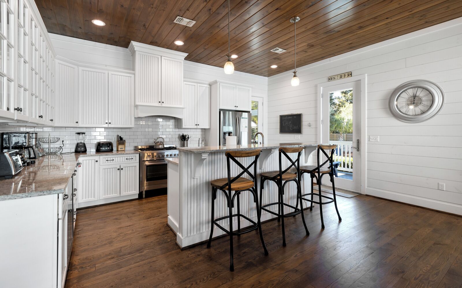 A spacious kitchen with white cabinetry, a central island with bar stools, wooden flooring, and a large window.