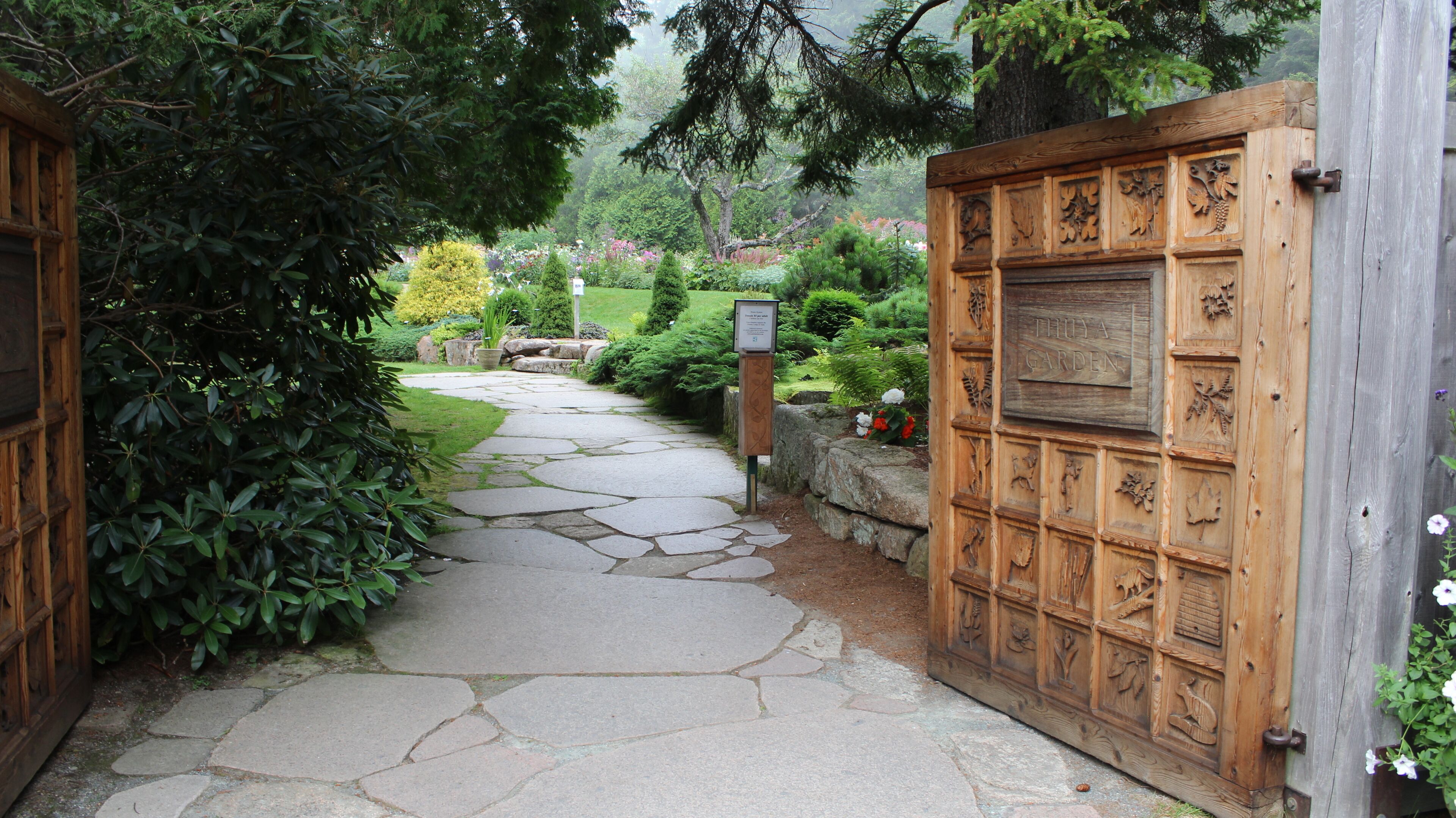 A stone pathway leading to a wooden gate with intricate carvings, surrounded by lush greenery and a sign indicating 'THUJA GARDEN'.
