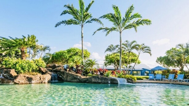 A clear pool with a waterfall feature, surrounded by palm trees and lounge chairs. - Poipu Beach Estates: Walk to Beach/Shops, Sleeps 8-16, AC, Pool, Fitness Club! (Koloa)