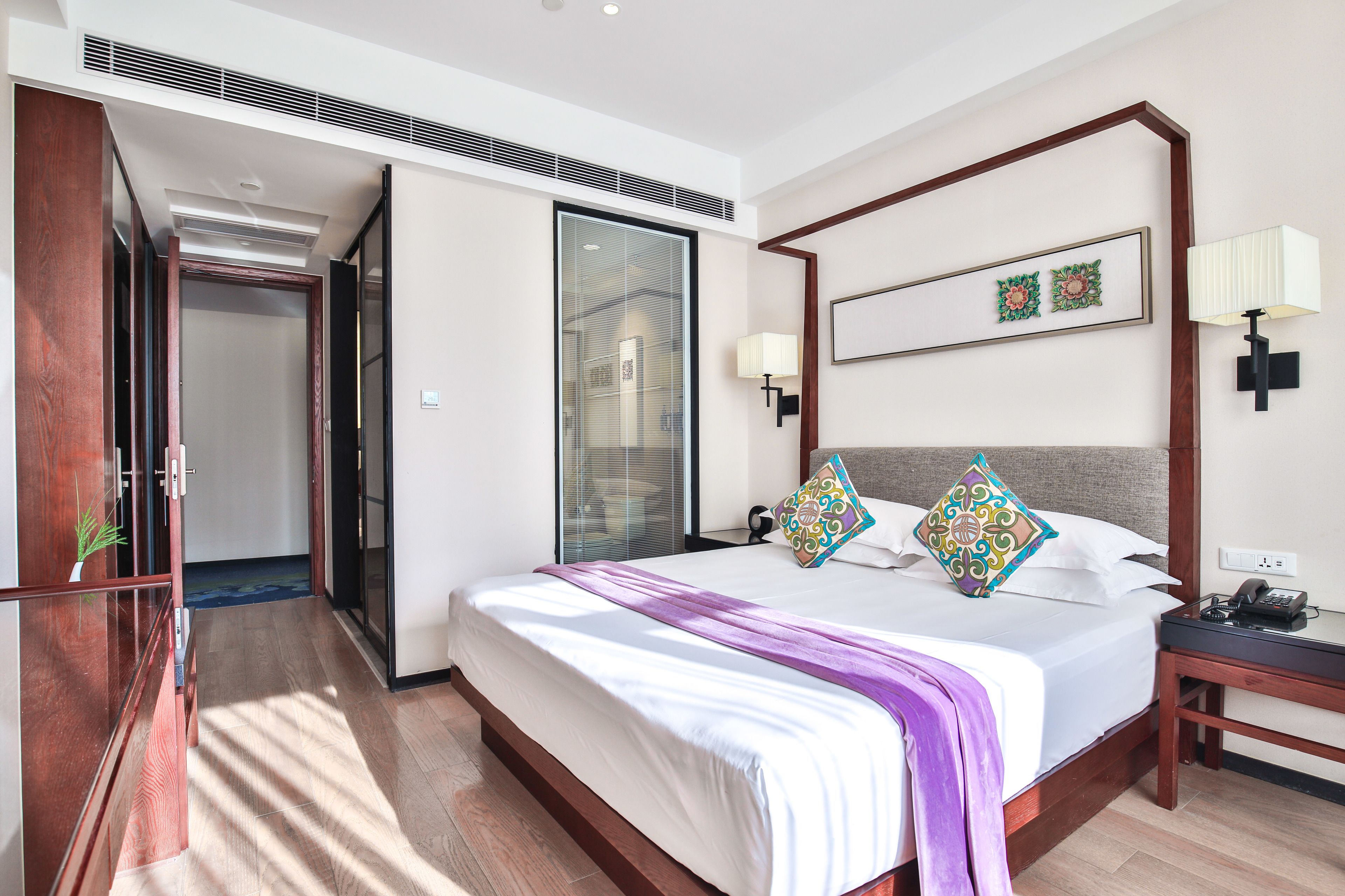 A hotel room with a large bed, a bedside table with a lamp, a wall-mounted artwork, and a sliding door leading to another room.