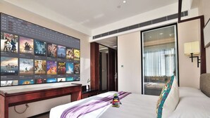 Ein Hotelzimmer mit einem großen Flachbildfernseher, der Filmplakate zeigt, einem Bett mit weißen Bettwäsche und bunten Kissen, einem Holztisch und einer Schiebetür, die auf einen Balkon führt.