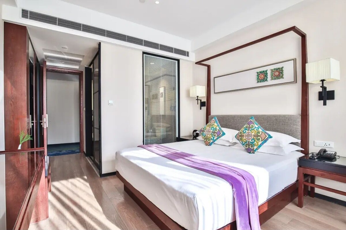 A hotel room with a large bed, a bedside table with a lamp, a wall-mounted artwork, and a sliding door leading to another room.