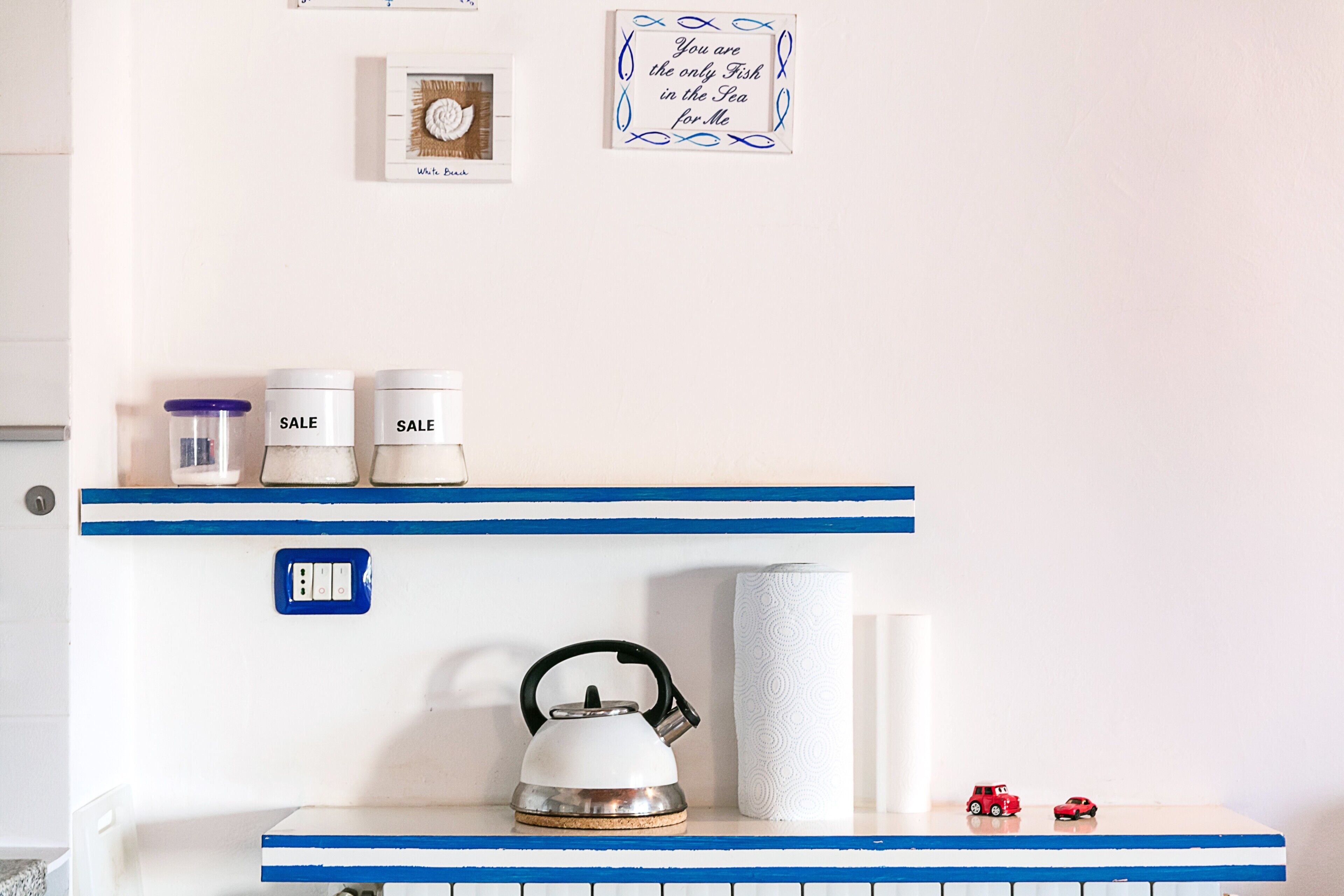 A kitchen with a kettle, two sale jars, a roll of paper towels, and two toy cars on a shelf.