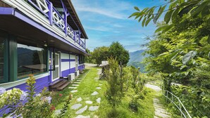 A house with a stone pathway leading to the entrance, surrounded by lush greenery and mountains in the background. - Julie's Garden (Ren'ai)