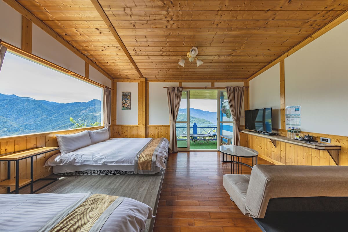 A room with wooden walls and ceiling, two beds, a sofa, a TV, and a window with a mountain view.