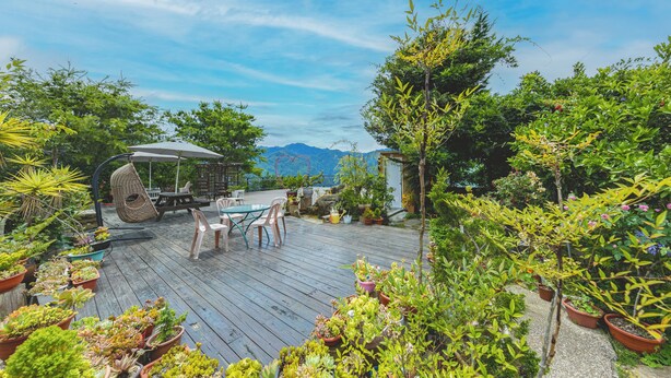A wooden deck with a table and chairs, surrounded by potted plants and a view of mountains. - Julie's Garden (Ren'ai)