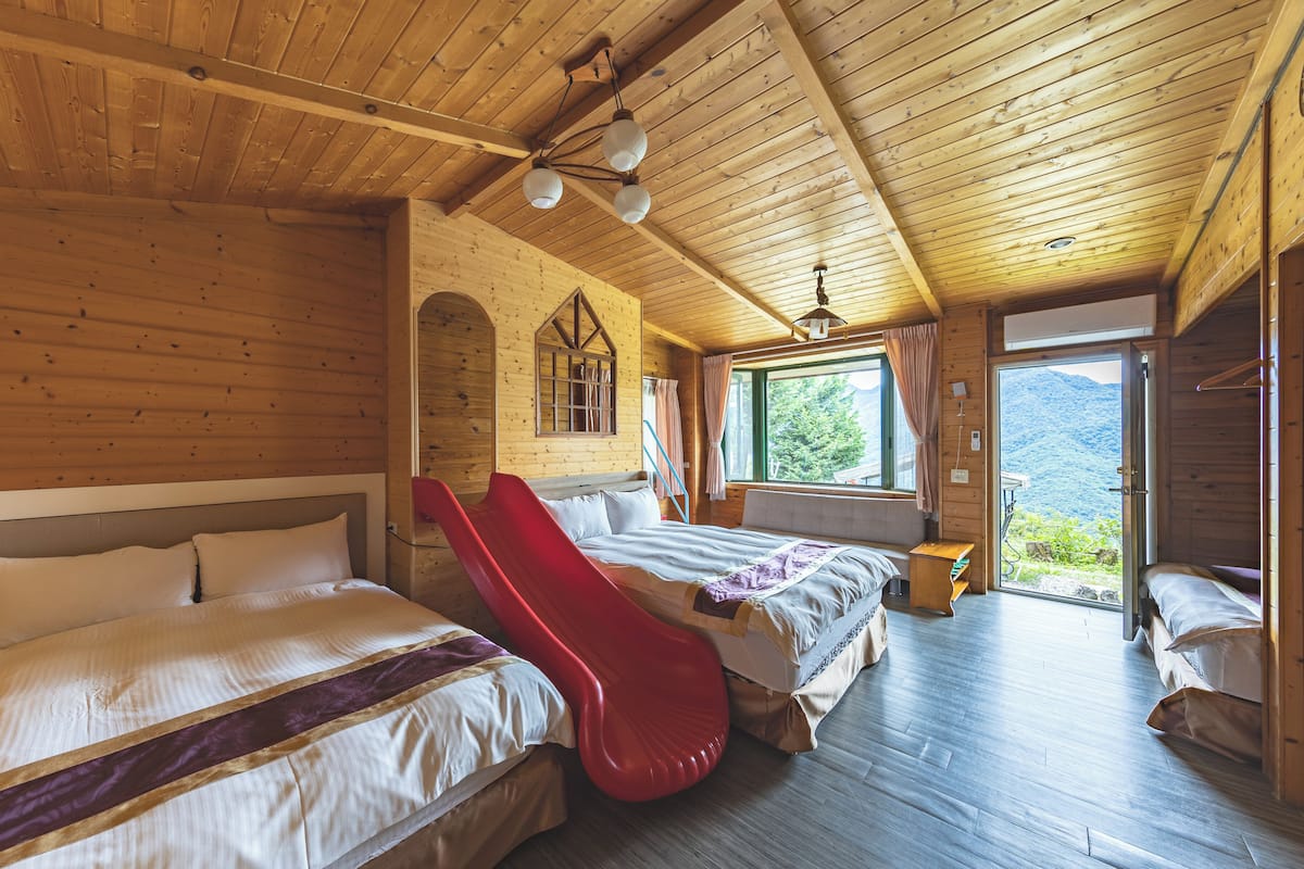 A wooden cabin room with two beds, a red slide, a window with curtains, and a view of mountains.