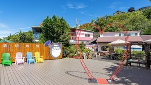A wooden deck with colorful Adirondack chairs, a red slide, and a building with a green roof in the background. - Julie's Garden (Ren'ai)